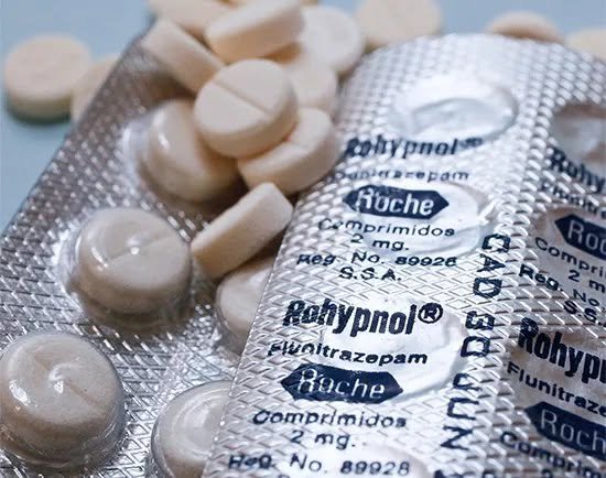 A hand holding a Rohypnol 2mg pill with a secure online pharmacy website displayed on a smartphone screen."